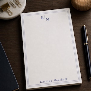 May include: A white notepad with a blue border and the initials "KM" at the top. The name "Katrina Marshall" is printed at the bottom in blue. A black pen, gold paper clips, and a lit candle are also visible.