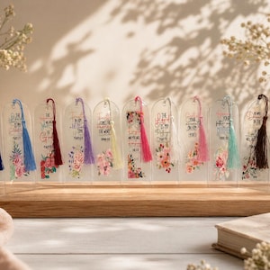 Peut inclure: Ensemble de marque-pages en acrylique transparent avec des motifs floraux colorés et des pompons. Chaque marque-page présente un verset biblique différent en texte noir. Les pompons sont de différentes couleurs, dont le bleu, le rose et le jaune. Les marque-pages sont sur un présentoir en bois.