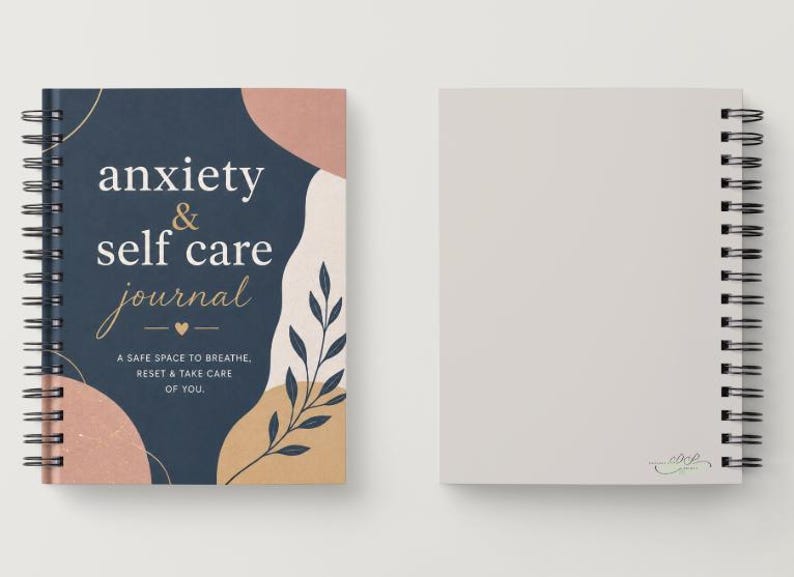 May include: A spiral-bound journal with a navy blue cover featuring the words "anxiety & self care journal" in white and gold script. The cover also includes abstract shapes in peach and beige, and a botanical illustration. The journal is open to a blank page.