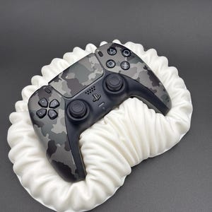 Peut inclure: Une manette de jeu vidéo à motif camouflage avec des boutons et des joysticks noirs repose sur une surface blanche et froissée. La manette a une base noire et un motif camouflage gris et vert sur le dessus.