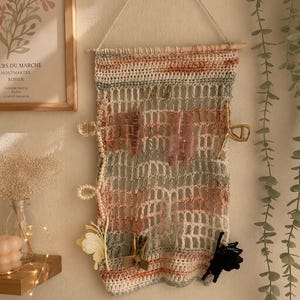 May include: A handmade, macrame wall organizer in shades of white, peach, and green, with a wooden dowel for hanging. The organizer holds various hair accessories, including pink claw clips and gold-toned hair clips. A decorative piece for home decor.