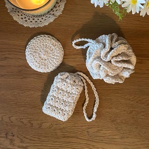 May include: A selection of handmade, cream-coloured crochet items. The set includes a round coaster, a loofah-style body scrubber, and a soap saver bag with a braided cord. The items are arranged on a wooden surface, suggesting a spa or bath theme.