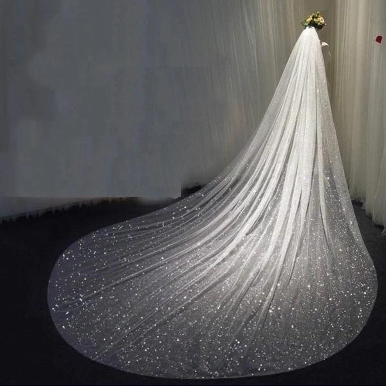Op de afbeelding: Een lange, witte tule bruids sluier met een sprankelend, verspreid glitter ontwerp. De sluier is over een oppervlak gelegd en heeft een kathedraallengte.