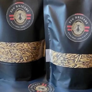 May include: Black bags of Egg Brigade product with a clear window showing the contents. The bags have a circular logo with text and an insect illustration. The bags are filled with a tan-colored product.
