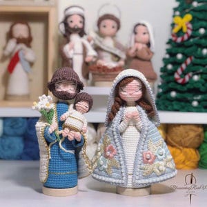 Könnte beinhalten: Gehäkelte Figuren von Josef, der das Jesuskind hält, und Maria in einem blau-weißen Kleid mit Blumenstickerei. Die Figuren stehen auf Holzsockeln.