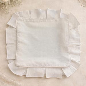 Puede incluir: Una almohada de lino blanco con borde con volantes, sobre un fondo neutro. La almohada tiene forma cuadrada con un borde contrastante. Estética suave y romántica, con detalles florales y de encaje.