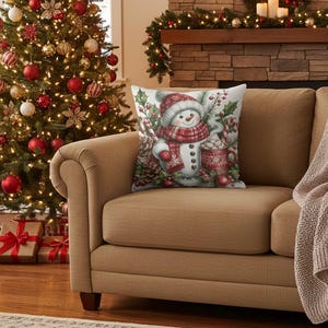 May include: A decorative Christmas pillow featuring a snowman wearing a red and white scarf and hat, holding a mug of hot cocoa. The pillow is on a tan couch in front of a Christmas tree and fireplace.