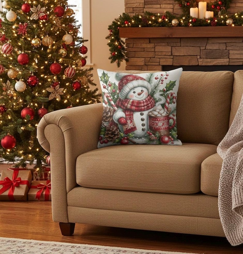 May include: A decorative Christmas pillow featuring a snowman wearing a red and white scarf and hat, holding a mug of hot cocoa. The pillow is on a tan couch in front of a Christmas tree and fireplace.