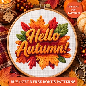 May include: A circular wooden embroidery hoop containing a cross-stitch design on white fabric. The design features the text Hello Autumn in yellow, surrounded by a cluster of orange, red, and green maple leaves. The hoop has a metal screw top.