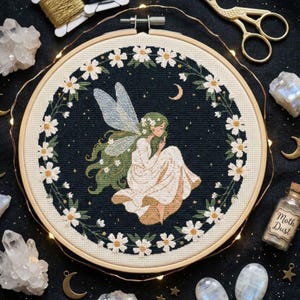 Puede incluir: Bordado de punto de cruz de un hada con cabello y alas verdes, vestida con un vestido blanco, rodeada por una corona floral. El fondo es azul oscuro con estrellas y una luna creciente. El bordado está en un aro de madera.