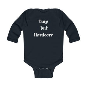 Puede incluir: Body de bebé azul marino con mangas largas. El texto "Tiny but Hardcore" está impreso en blanco, con una fuente estilizada en la parte posterior. El body tiene tres cierres de botón a presión en la parte inferior.