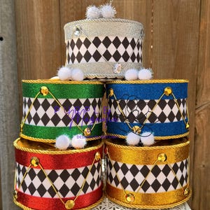 May include: A stack of five cylindrical drum-shaped containers featuring a black and white diamond pattern. The drums have glittery bands in silver, green, blue, red, and gold, accented with gold or silver rope trim, small white pom-poms, and gems.