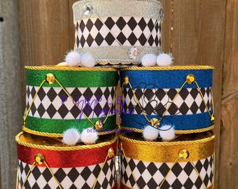 May include: A stack of five cylindrical drum-shaped containers featuring a black and white diamond pattern. The drums have glittery bands in silver, green, blue, red, and gold, accented with gold or silver rope trim, small white pom-poms, and gems.