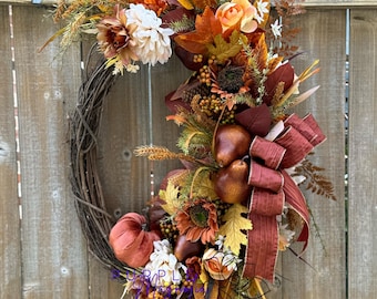 May include: A fall-themed wreath with a variety of decorative elements. It features a grapevine base adorned with faux pumpkins, pears, sunflowers, and colorful autumn leaves in shades of orange, brown, and yellow. A burgundy ribbon adds a finishing touch.