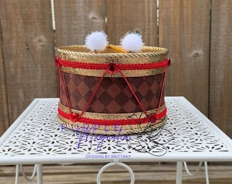 May include: A decorative drum with a brown and gold diamond pattern. The drum has red and gold trim, with white pom-poms and drumsticks. The drum is sitting on a white metal table.