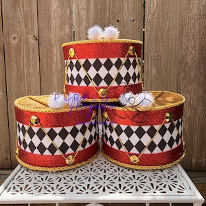 May include: Three cylindrical drum-shaped containers featuring a red glitter border, a black and white diamond pattern, and gold rope trim. Each container is topped with a gold lid and a white pom-pom. The text Purple Magnolia is visible.
