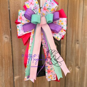 May include: A decorative bow with multiple layers of colorful ribbons. The bow features a floral print, solid red, purple, mint green, and pink ribbons. The bow is hanging on a wooden fence. The text "Purple Magnolia" is visible.