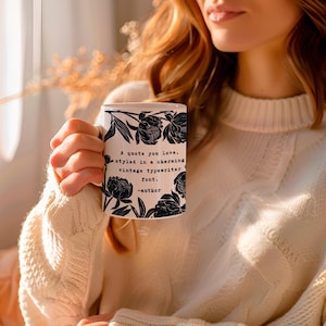 May include: A white ceramic mug featuring a black floral pattern of peonies surrounding the printed text: A quote you love, styled in a charming vintage typewriter font. -author. The mug has a cylindrical shape and a handle on the right side.