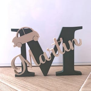 Puede incluir: Una letra M de madera pintada de color verde oscuro con el nombre "Martin" escrito en color beige en la letra. Un pequeño coche beige está fijado en la parte superior de la letra.