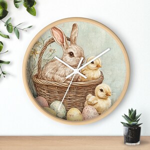 Könnte beinhalten: Runde Wanduhr aus Holz mit Ostermotiv. Das Zifferblatt zeigt einen Hasen und zwei Küken in einem Weidenkorb, umgeben von bemalten Eiern. Die Uhr hat einen hellgrünen Hintergrund und einen Rahmen aus Naturholz.
