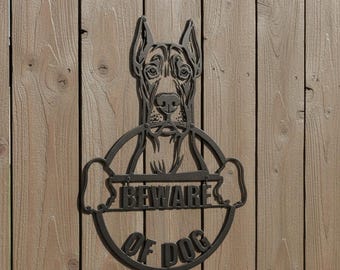 May include: A black metal sign featuring a Great Dane dog head and the words "BEWARE OF DOG" in a circular design. The sign is mounted on a wooden fence with vertical planks.