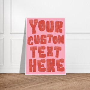 Könnte beinhalten: Ein gerahmtes rosa Poster mit den Worten "YOUR CUSTOM TEXT HERE" in großen, roten Buchstaben. Das Poster lehnt an einer weißen Wand auf einem Holzboden.