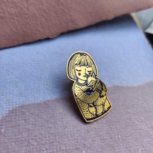 Peut inclure: Une broche en métal doré émaillé représentant l'illustration stylisée d'une personne au carré portant un pull texturé et tenant un petit renard. Le motif présente des contours noirs, des touches rouges sur les joues et une finition dorée.