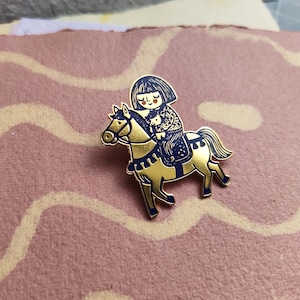 Peut inclure: Une broche en émail et métal doré représentant une illustration stylisée d'une personne aux cheveux foncés montant un cheval. Le cheval est fini en doré avec des accents bleu foncé sur la crinière, la queue, les sabots et la selle.