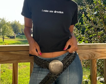 May include: A black cropped t-shirt with the text "i miss one direction" in white embroidery. The shirt is being worn by a person with a braided hairstyle and a black braided belt with a silver buckle.