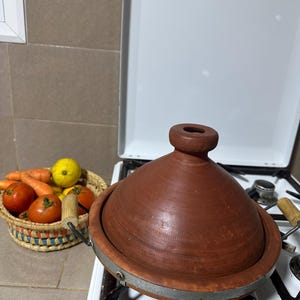 May include: A brown earthenware tagine pot with a conical lid sits on a white stovetop. A woven basket filled with tomatoes, carrots, and lemons is to the left. The tagine is a traditional North African cooking vessel.