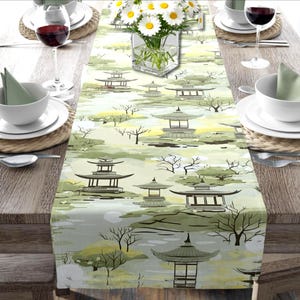 May include: A table runner with a Japanese garden design. The runner features pagodas, trees, and water elements in shades of green, gray, and yellow. The table is set with white dishes, wine glasses, and a vase of daisies.