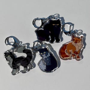 Op de afbeelding: Vier kat-thema sleutelhangers met zilverkleurige sluitingen. De katten zijn in verschillende poses en kleuren, waaronder zwart, grijs en oranje. De sleutelhangers zijn gemaakt van een transparant materiaal, waardoor de kattenontwerpen zichtbaar zijn.