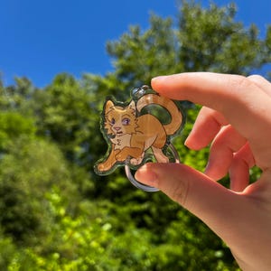 May include: A clear acrylic keychain featuring a cartoon cat with orange and white fur, purple eyes, and a black nose. The cat is in a walking pose with its tail curled. The keychain has a silver metal ring.