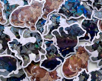 May include: A collection of cat stickers with a holographic, geometric design. The stickers feature various cat poses in shades of brown, blue, and gray. The stickers have a white border and are scattered across a white surface.