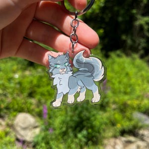 May include: A clear acrylic keychain featuring a stylized gray cat with blue and white accents. The cat has green eyes and a fluffy tail. The keychain is attached to a silver metal chain and ring.