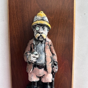 May include: A wall-mounted plaque featuring a three-dimensional figure of a man wearing a yellow pith helmet, a monocle, and a tan suit. The figure holds a glass and stands on a base labeled GIN, all mounted on a rectangular dark wood board.