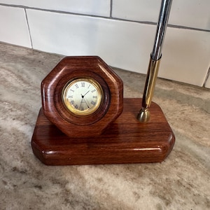 May include: A desk accessory featuring a polished dark wood base with an octagonal clock housing. The clock has a gold-tone bezel, a white face with black Roman numerals, and thin black hands. A metallic pen holder stands upright on the base.