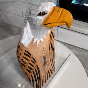 May include: A hand-carved wooden light switch cover featuring a bald eagle design. The eagle has a white head, a bright yellow beak, and a natural wood-toned body with black painted feather details. A rectangular cutout is centered for the switch.