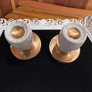 May include: Two cylindrical candle holders featuring a speckled cream and brown exterior finish. Each holder has a smooth, metallic gold interior and a matching circular gold base, providing a contrast between the textured top and the polished bottom.
