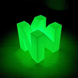 May include: A green glowing plastic sculpture of the Nintendo 64 logo. The logo is made up of three interlocking blocks that form the letter "N".