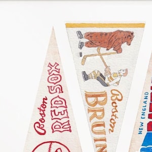 Puede incluir: Tres banderines vintage con logotipos y texto de equipos. Los banderines son blancos con texto y gr&aacute;ficos rojos, marrones y azules. Los banderines presentan los logotipos de los Boston Red Sox, Boston Bruins y New England Patriots. Los banderines tienen forma triangular.