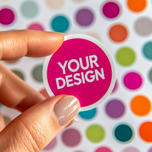 May include: A magenta circular sticker with the text "YOUR DESIGN" in white. The sticker is held by a hand against a backdrop of multi-coloured polka dots. The sticker is a promotional item.