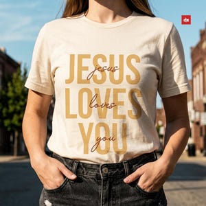 Puede incluir: Una camiseta color crema con el mensaje "JESUS loves YOU" impreso en negrita, en oro. La camiseta es de manga corta y la lleva una persona con las manos en los bolsillos. El fondo muestra una escena de calle borrosa.