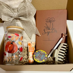 Puede incluir: Una caja de regalo con un surtido temático de otoño. Incluye un vaso de vidrio con un diseño de calabaza, un diario con el nombre "Sophia", un bolígrafo, una pinza para el pelo, crema de manos y un recipiente decorativo.