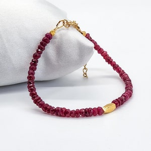 May include: A delicate bracelet featuring small, faceted ruby beads strung together. The bracelet includes a gold-colored clasp and a gold-colored, elongated bead as a central accent. The jewelry is displayed against a white background.