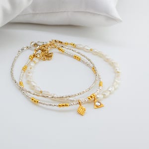 May include: Three delicate bracelets with gold and silver beads and white pearls. The bracelets have small gold charms in the shape of a diamond and a triangle.