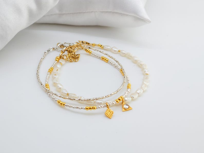 May include: Three delicate bracelets with gold and silver beads and white pearls. The bracelets have small gold charms in the shape of a diamond and a triangle.
