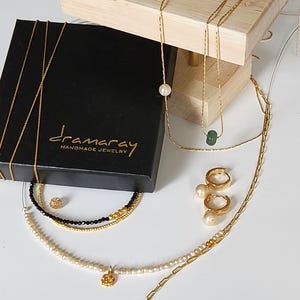 May include: A black box with the text "dramarray handmade jewelry" on it. The box is open and contains a variety of gold necklaces and earrings with pearls and a green stone.