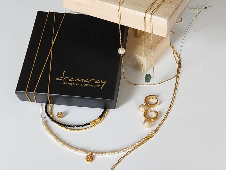 May include: A black box with the text "dramarray handmade jewelry" on it. The box is open and contains a variety of gold necklaces and earrings with pearls and a green stone.