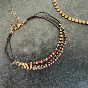 May include: A gold-coloured bracelet with three strands of small, rectangular beads. The bracelet features red and blue beads interspersed among the gold beads. A gold-coloured chain and clasp are visible. A gold-coloured necklace is in the background.